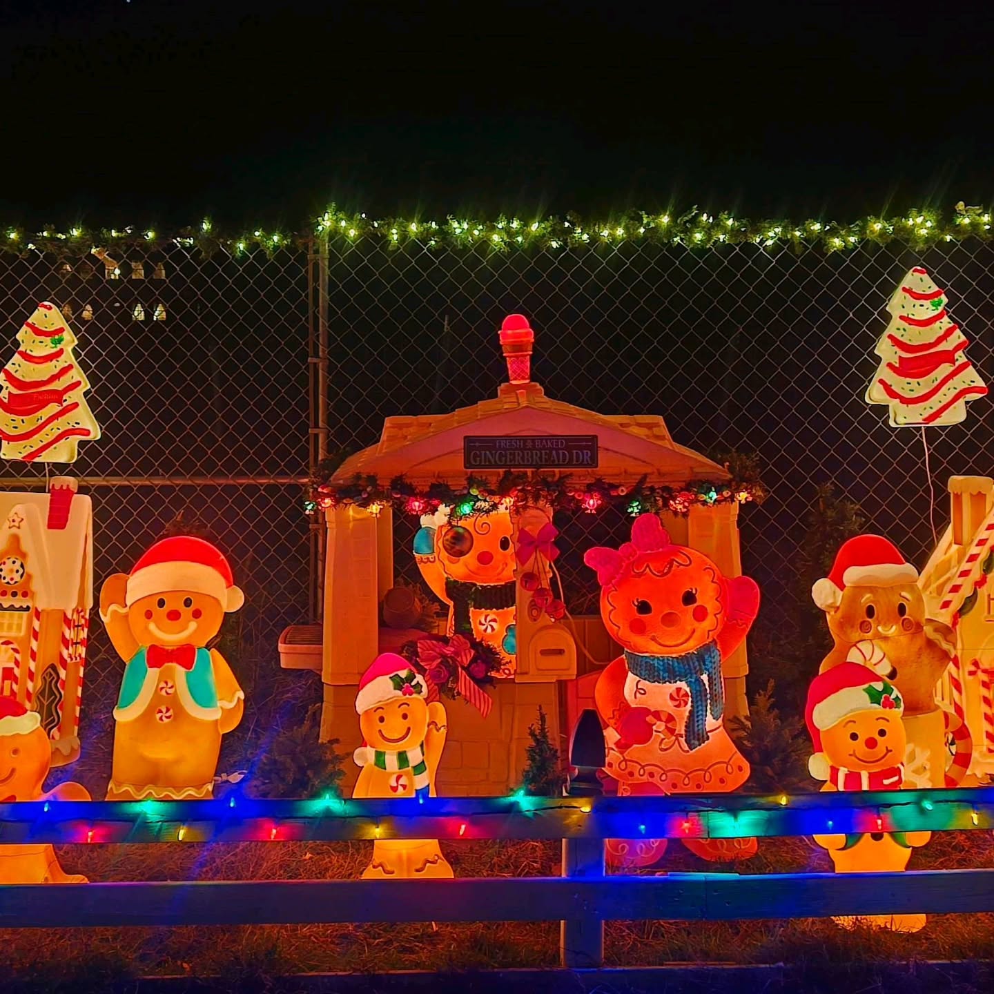 Illuminated collection of retro gingerbread man and woman blow mold figures standing in front of a decorated mini gingerbread cottage at night.