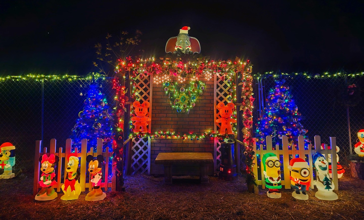 An illuminated photo bench beneath an evergreen arch flanked by glowing Christmas trees and character blow molds including Mickey, Minnie, Olaf, and Minions.