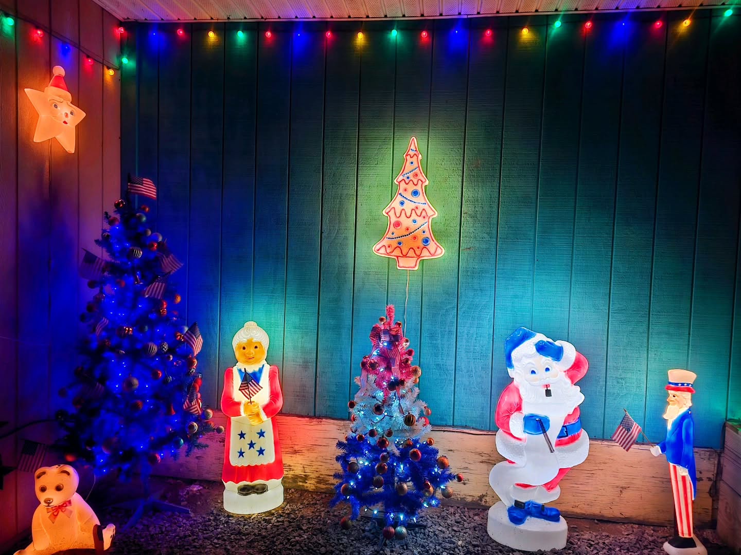 Indoor holiday display with illuminated blow mold figures of Uncle Sam, Santa, and Mrs. Claus in American flag attire beside miniature flag-decorated Christmas trees.
