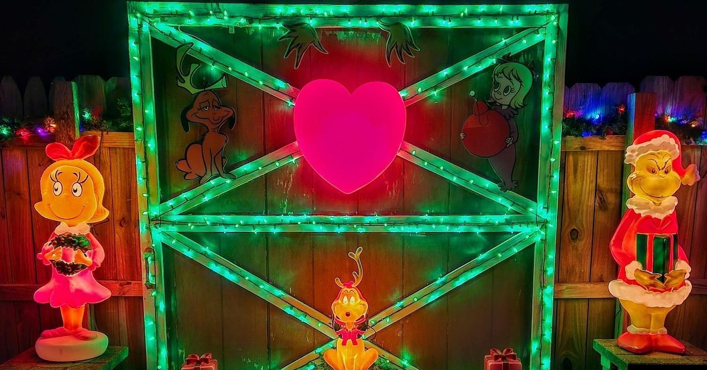 A rustic wooden gate decorated with green string lights and a large glowing heart in the center. Blow molds of the Grinch, Cindy Lou Who, and Max the dog surround the gate in a festive holiday display.